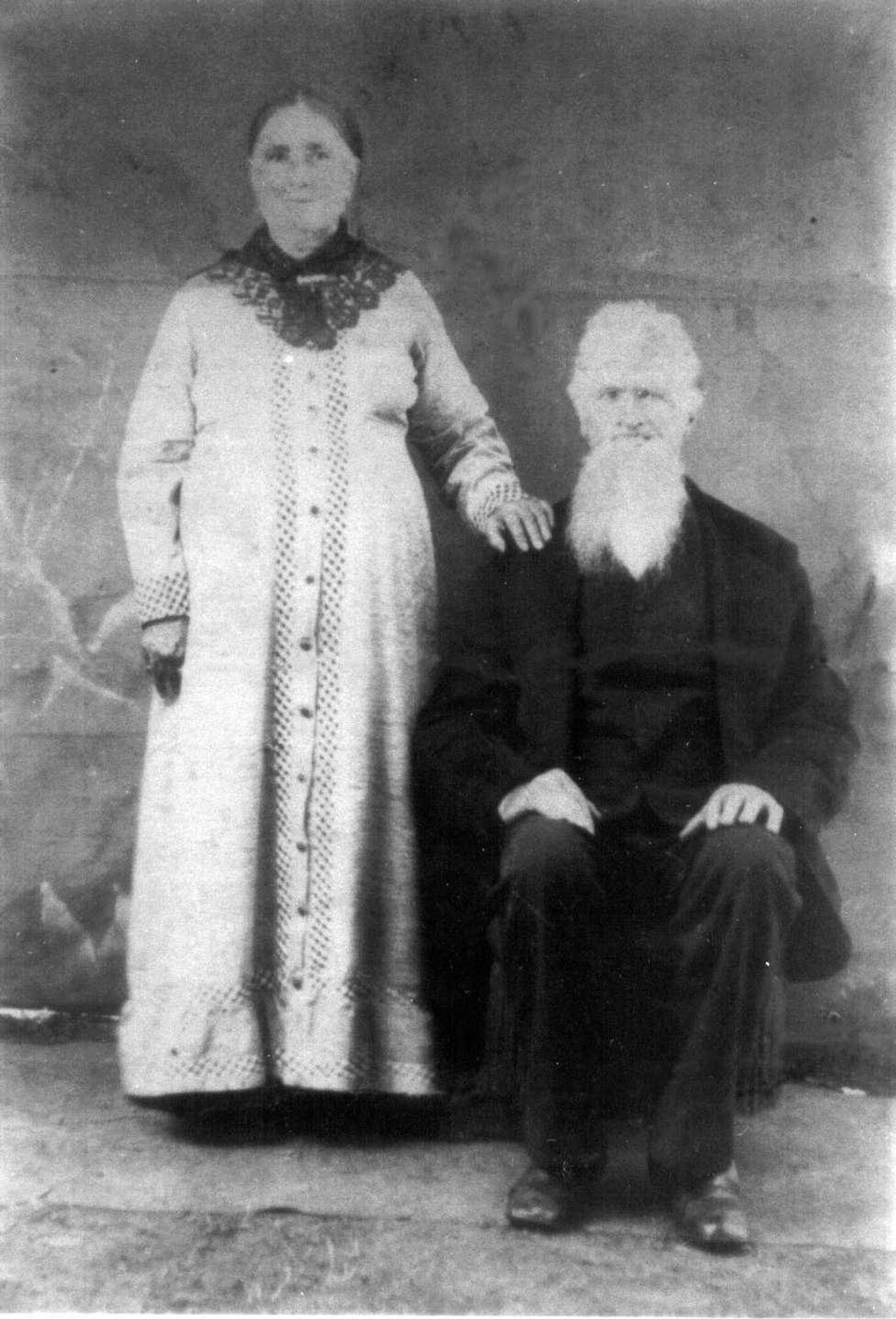 Woman dressed in white dress standing next to man who is sitting dressed in black overcoat.