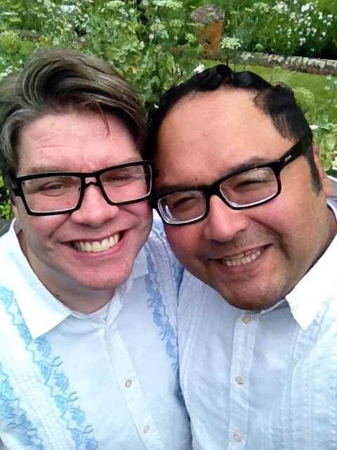 Two men dressed in blue shirts and black glasses smiling