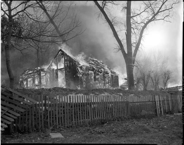 House ablaze with a picket fence in front and trees and smoke surrounding it.