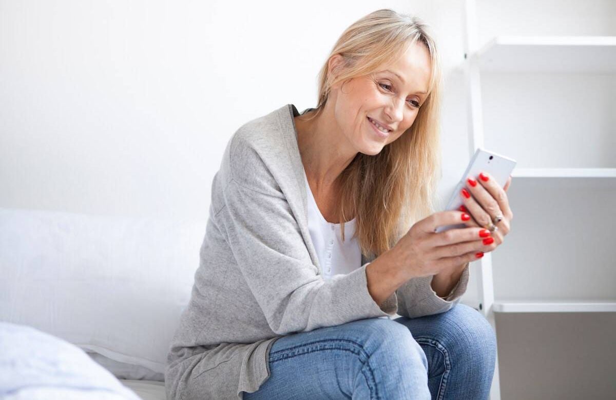 Mature woman with smart phone sitting on bed