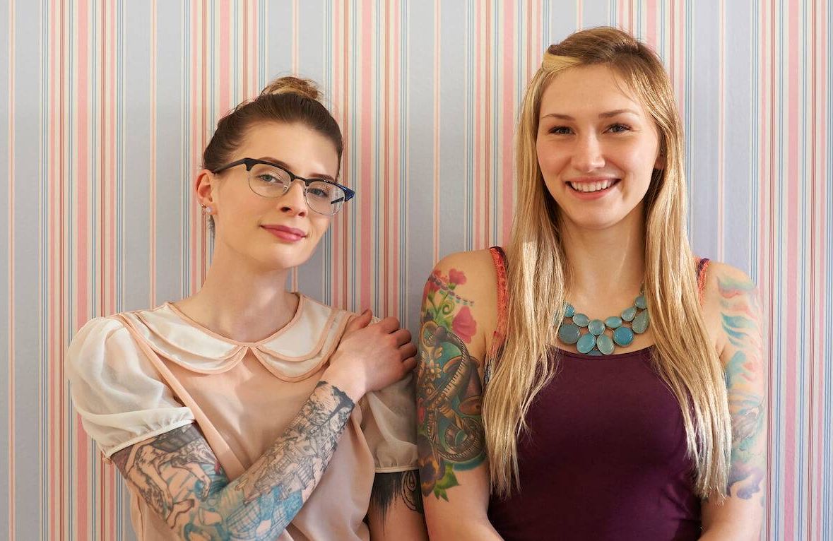 Two women in their 20s with tattooed arms stand against a vintage style wallpaper and smile.