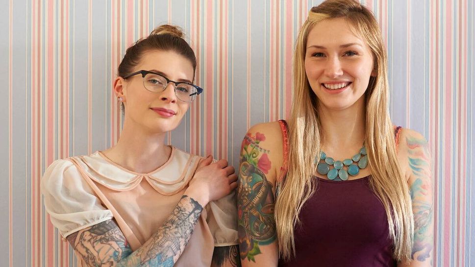 Two women in their 20s with tattooed arms stand against a vintage style wallpaper and smile.