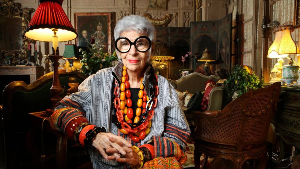 Fashion icon Iris Apfel seated, wearing famous black round glasses and many accessories