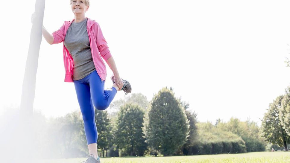 Senior woman leaning on tree trunk doing stretching exercises in a park