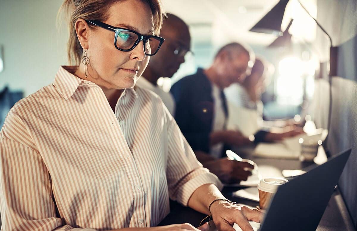 Female designer working on a laptop while sitting in a row with coworkers at a table in a modern office