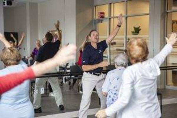 A man leads a group of older adults in an exercise class. They all have their arms raised and are moving with him. 