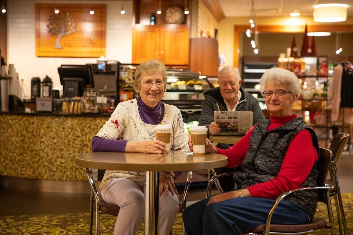 Two women and a man sit at a restaurant table with paper coffee cups. They are smiling at the camera.