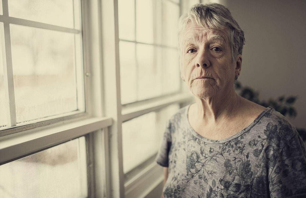 older woman standing near a window, looking sad