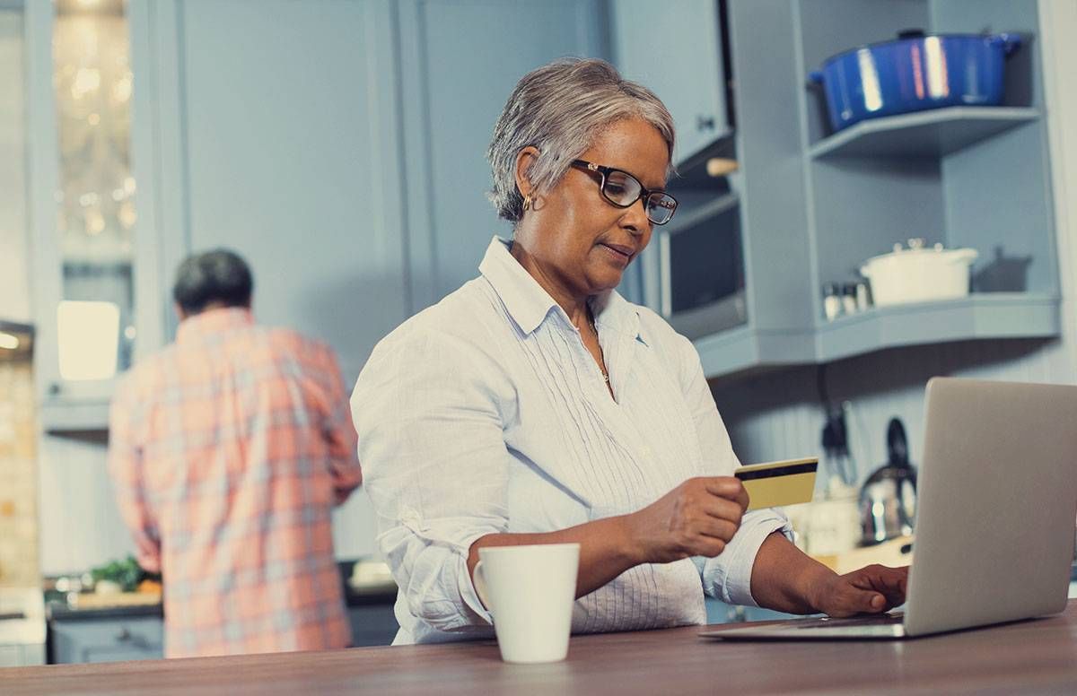 a retired couple in their kitchen; husband is preparing something and the woman is entering credit card information on a computer