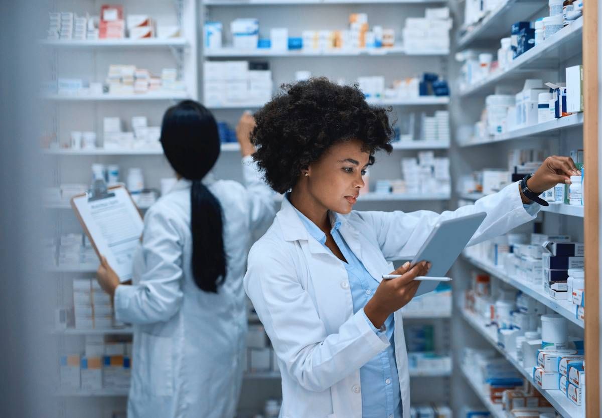 A person looking at a tablet in front of a large shelf of medications. She appears to be within a pharmacy.