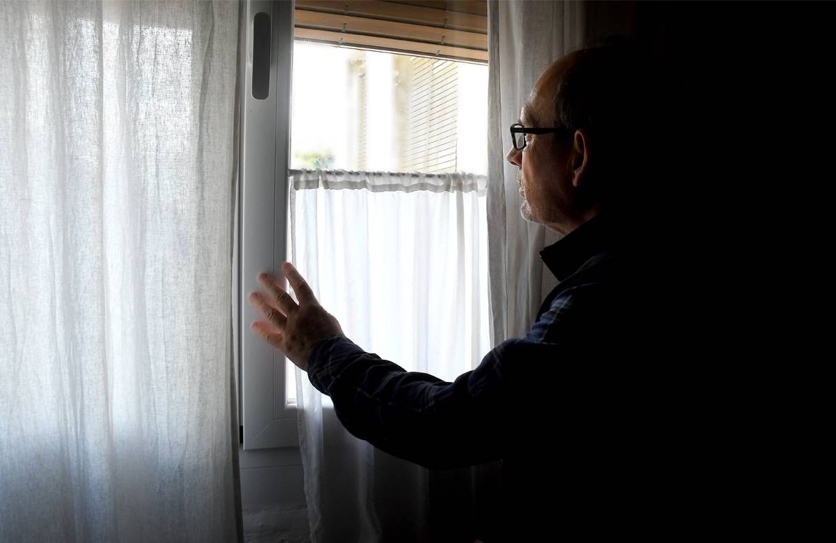 man looking out of the window of his dark home