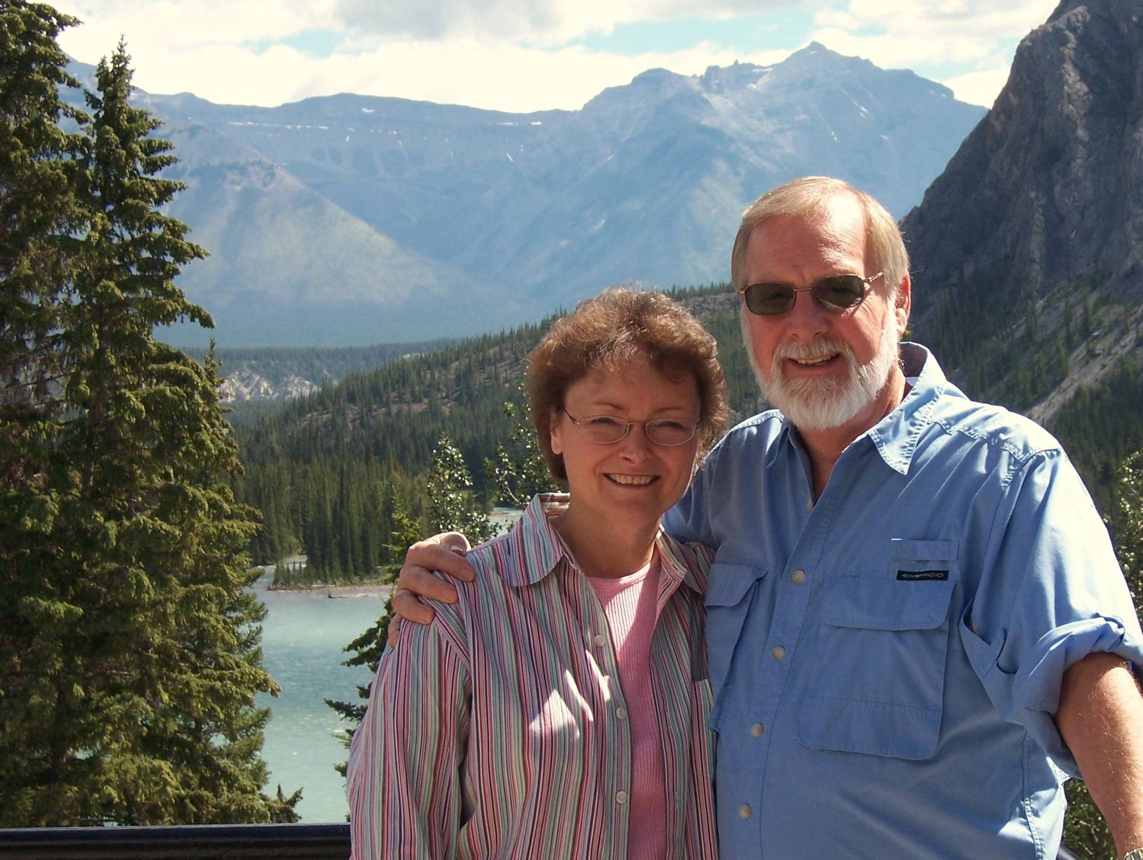 David and Linda stand in front of a picturesque scene of a lake and mountains.