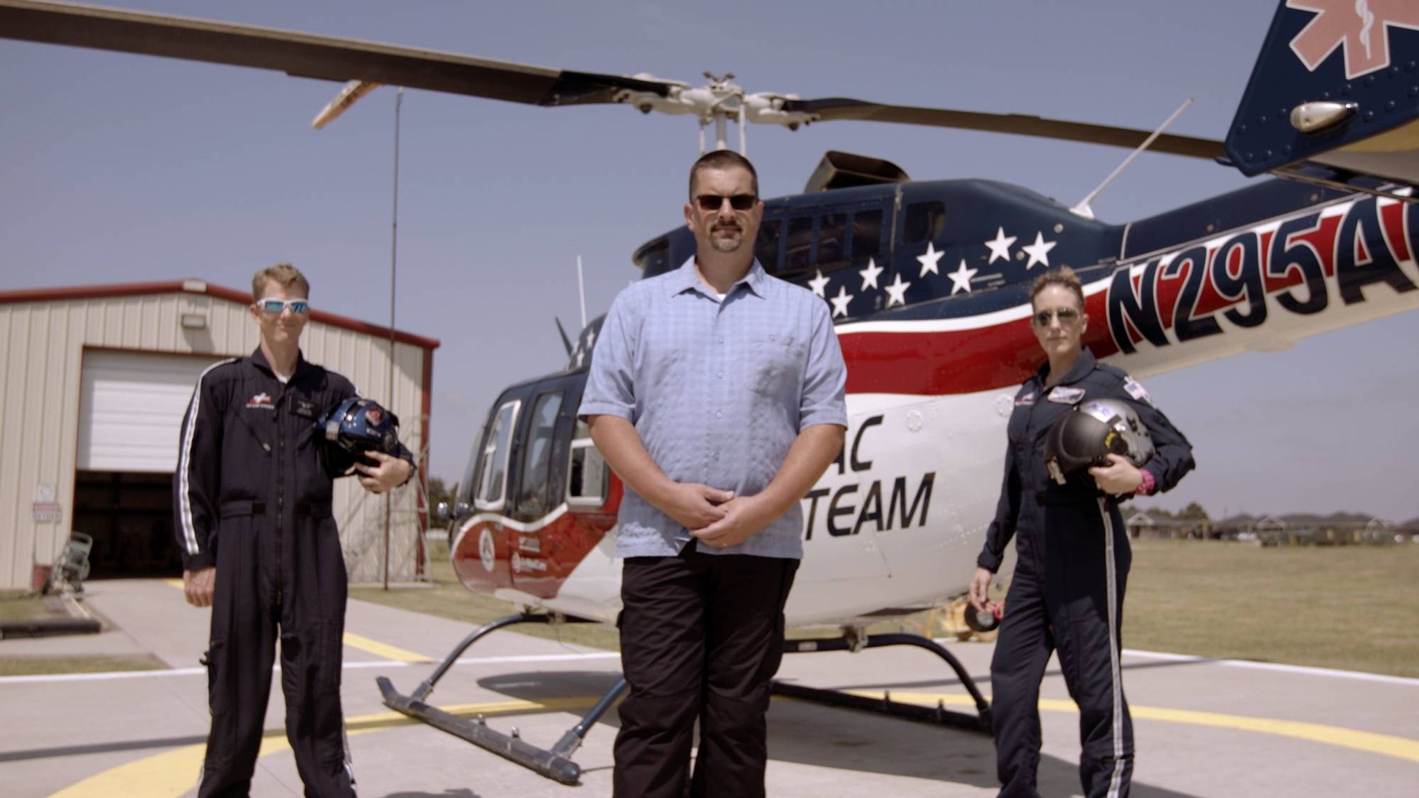 Three men standing on a helicopter tarmac with a helicopter behind them. The man in the center is wearing a blue shirt and sunglasses. The men to either side are in uniform.