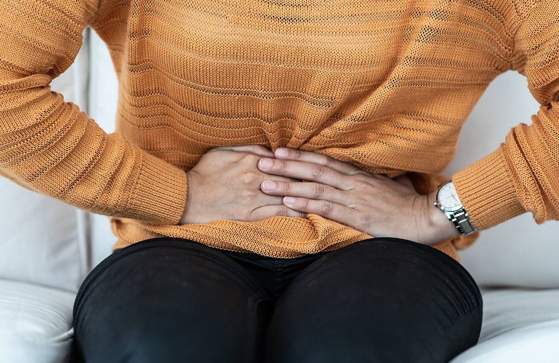 Closeup of a woman holding her bladder in pain sitting on couch, UTI, Next Avenue