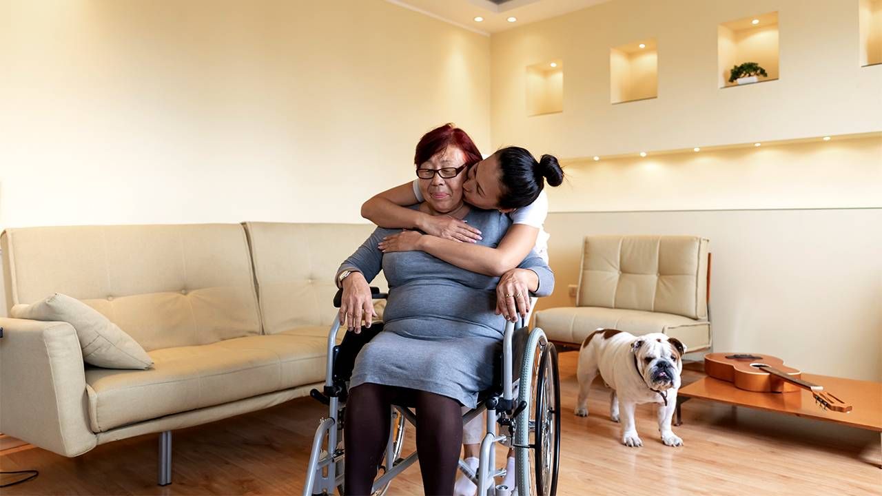 Older woman in wheelchair being hugged by her caregiving daughter, caregiver, caregivers, financial strain, Next Avenue