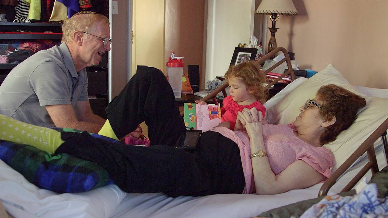 Toddler sitting on bed with older woman who is laying down next to an older man, caregiver, caregiving, Next Avenue