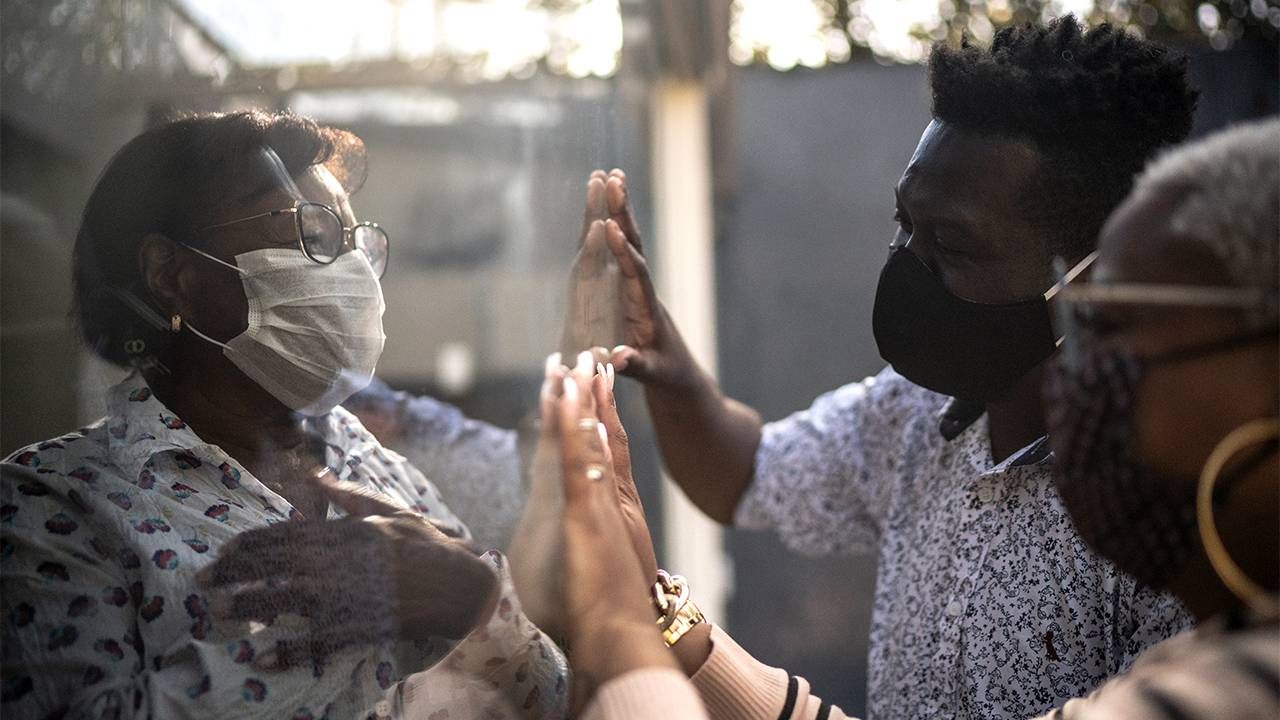 Older woman wearing face mask waving to family members through a window, aging, pandemic, Next Avenue