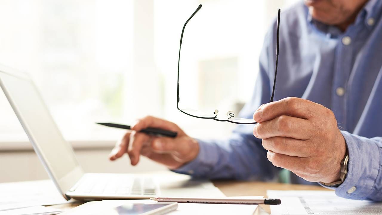 An older man holding his glasses while typing on a computer. Credit score, credit report, Next Avenue