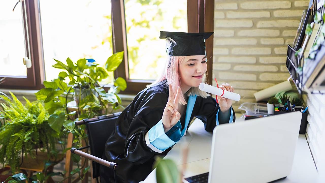 A young college graduate wearing a cap and gown for online graduation ceremony. Zoom, Next Avenue
