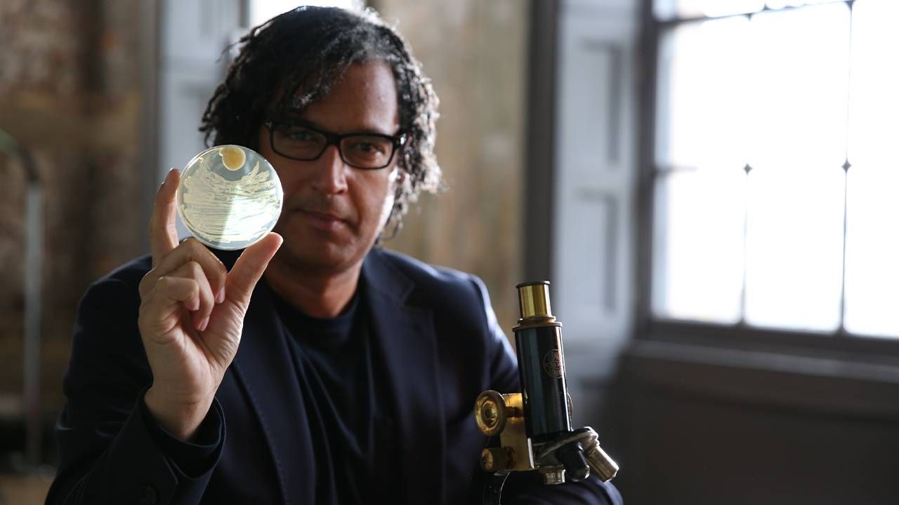 A man in front of a microscope holding up a petri dish. Extra life, PBS, Next Avenue