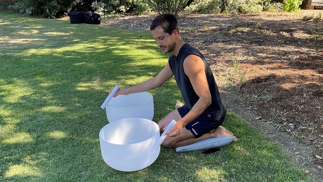 A man kneeling on a pillow playing the singing bowls for a sound bath. Next Avenue, stress