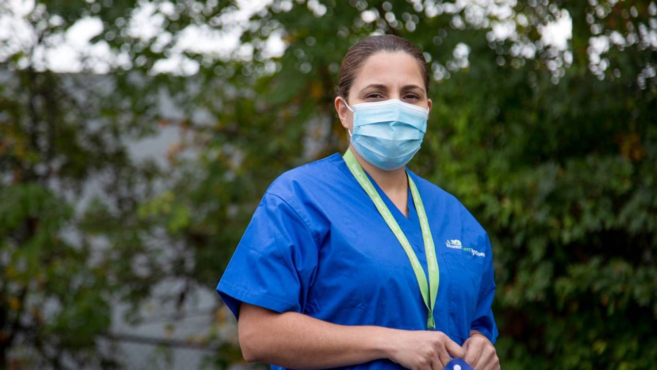 A woman standing in a parking lot wearing scrubs and a face mask. Next Avenue, paid caregivers, CNAs, caregiving work