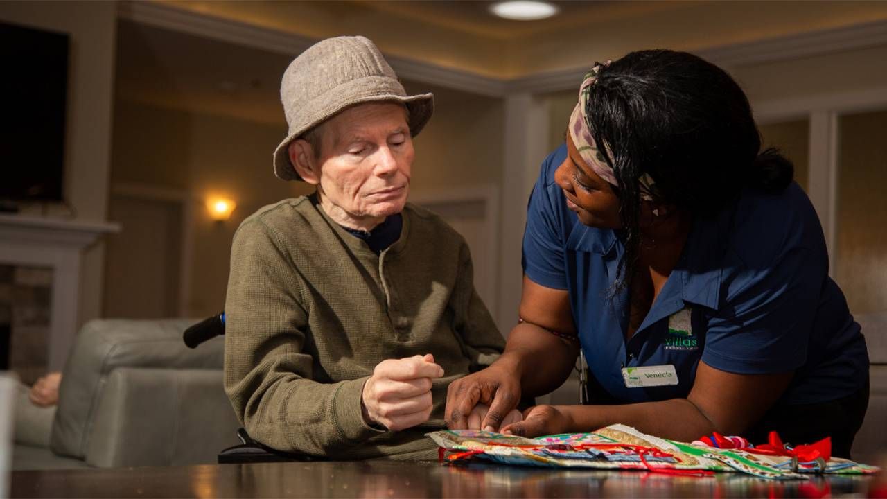 A woman wearing scrubs helps an older man in a wheelchair. Next Avenue, paid caregivers, CNAs, caregiving