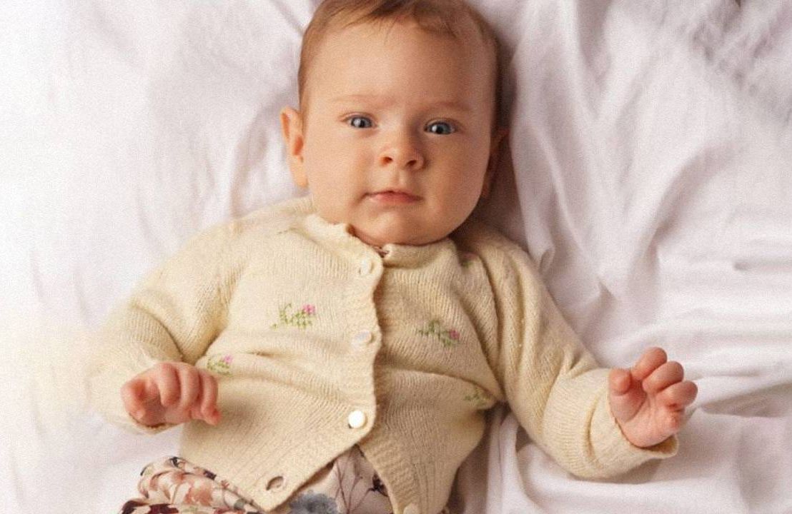 A baby wearing a knitted sweater laying on a white sheet. Next Avenue, family heirloom, sweater