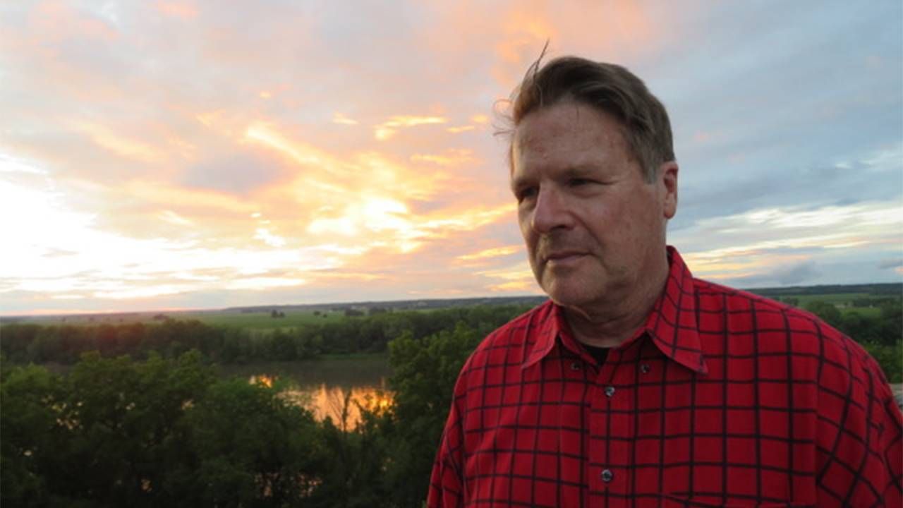 A man wearing a checked shirt standing in front of a sunset. Next Avenue