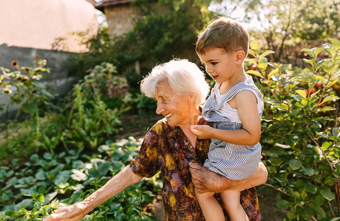 A woman in her old age holding a young grandchild showing him her garden, vitality, longevity, Next Avenue