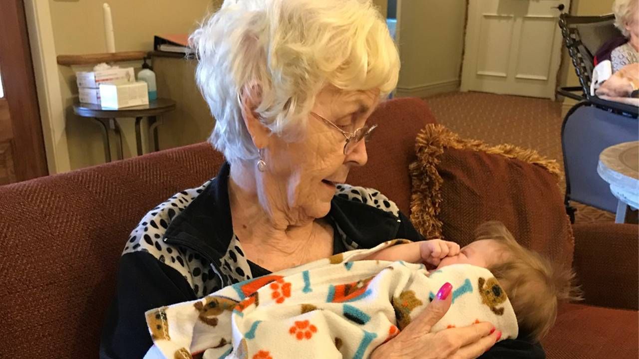 An older woman sitting in a chair holding an infant wrapped in blanket. Next Avenue, pandemic change nursing homes