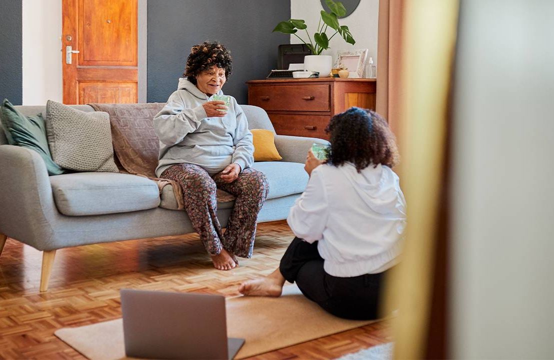 An older adult sitting on the couch with talking with adult daughter. Next Avenue, Parent's Sedentary Lifestyle