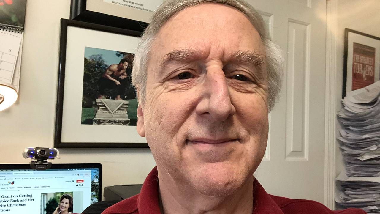 A man smiling at the camera inside a busy home office. Next Avenue