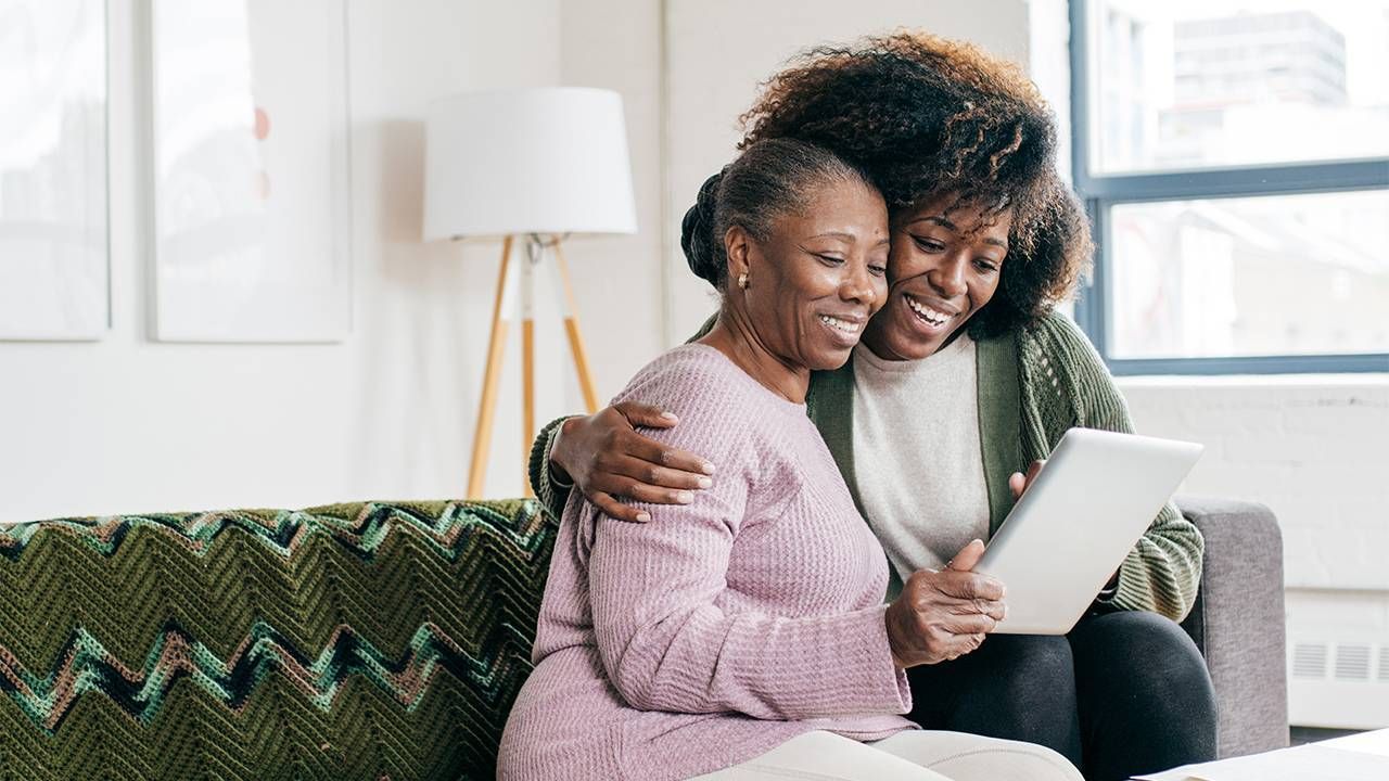 A younger woman caregiver embracing an older adult and holding an ipad. Next Avenue