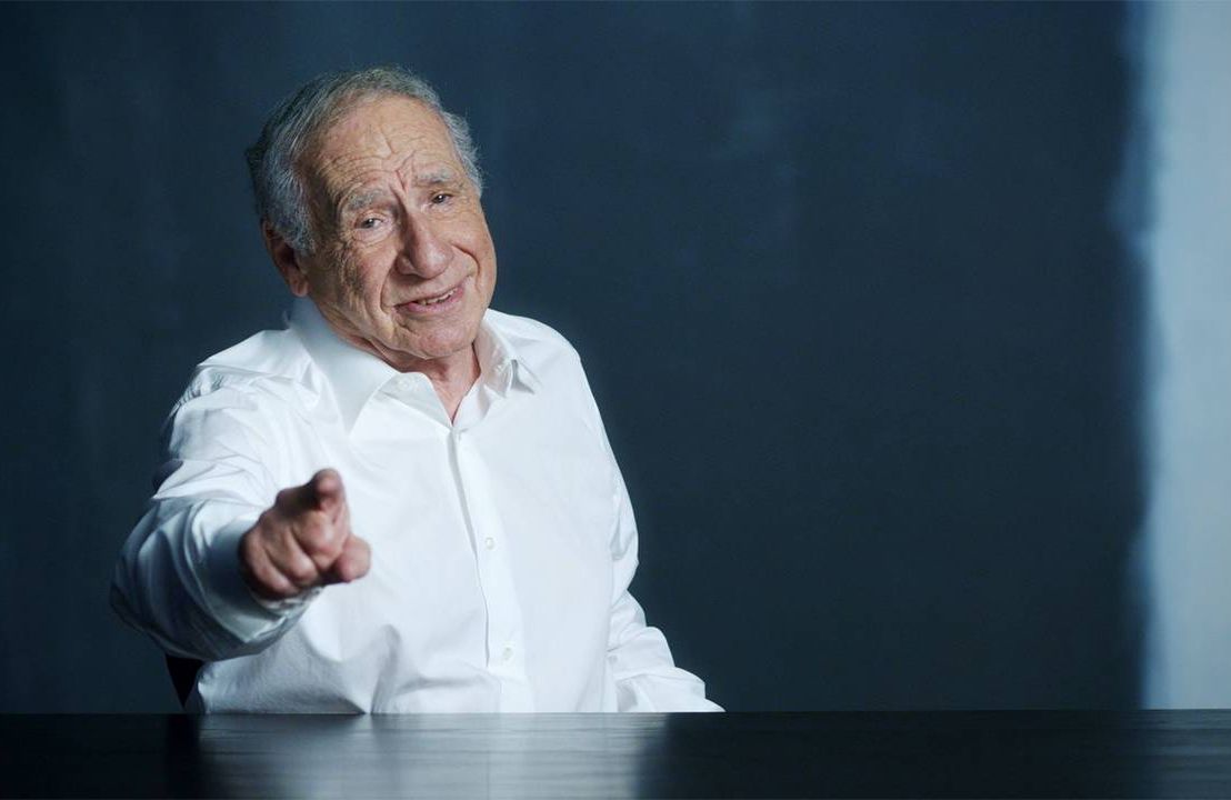 Mel Brooks wearing a white button up smiling and pointing at the camera. Next Avenue
