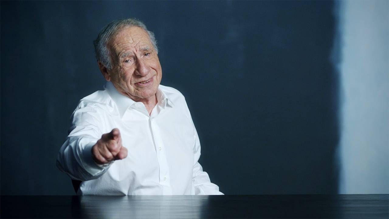 Mel Brooks wearing a white button up smiling and pointing at the camera. Next Avenue