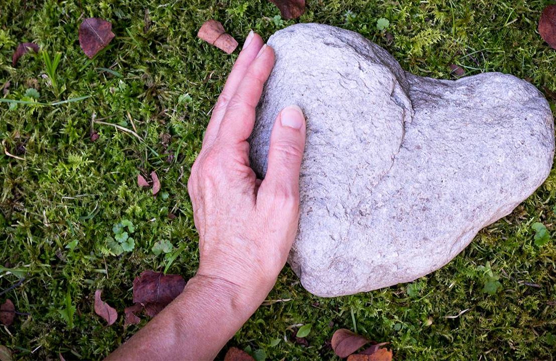 A person's hand touching a heart shaped rock in place of a tombstone in a green burial. Next Avenue,