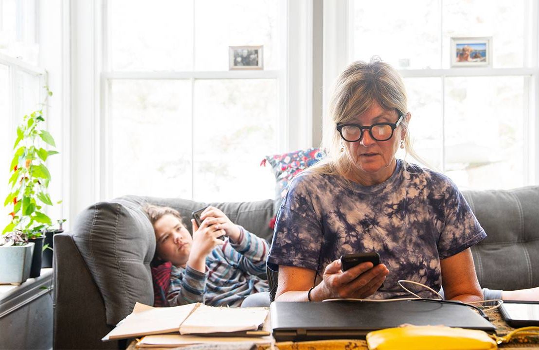 A mother looking at her cell phone while working from home with her child laying on the couch. Next Avenue, work life balance
