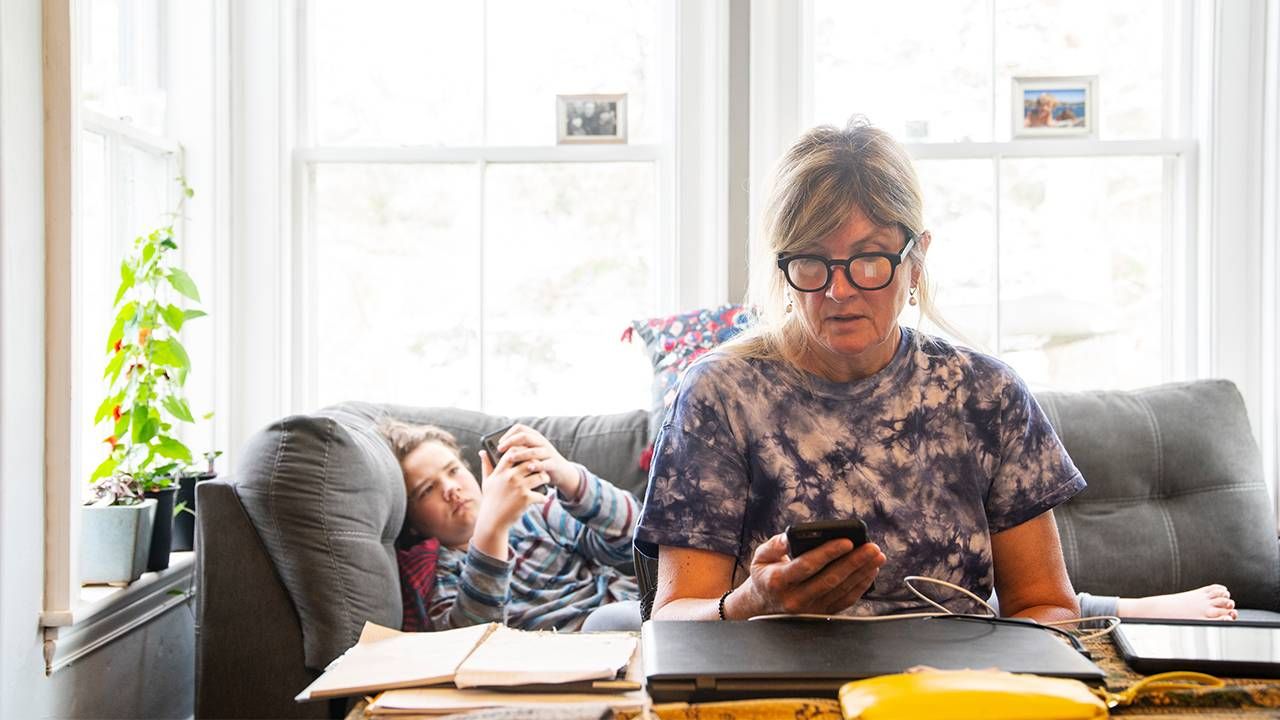 A mother looking at her cell phone while working from home with her child laying on the couch. Next Avenue, work life balance