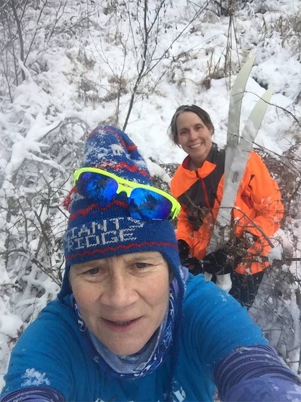 Tow older adults walking through thick woods in the snow, holding their skis. Next Avenue, skiing safely middle age