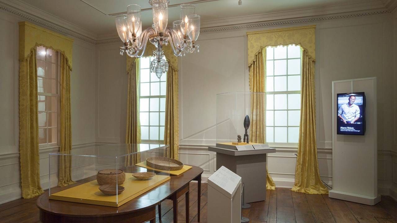 A living room with gold colored window curtains and woven baskets on display. Next Avenue, period rooms, MIA