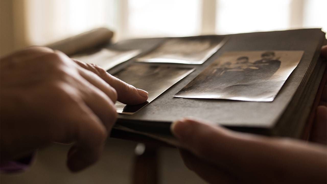 A close up shot of an old photo album and two people paging through the photos. Next Avenue, looking back, reminiscing about the past