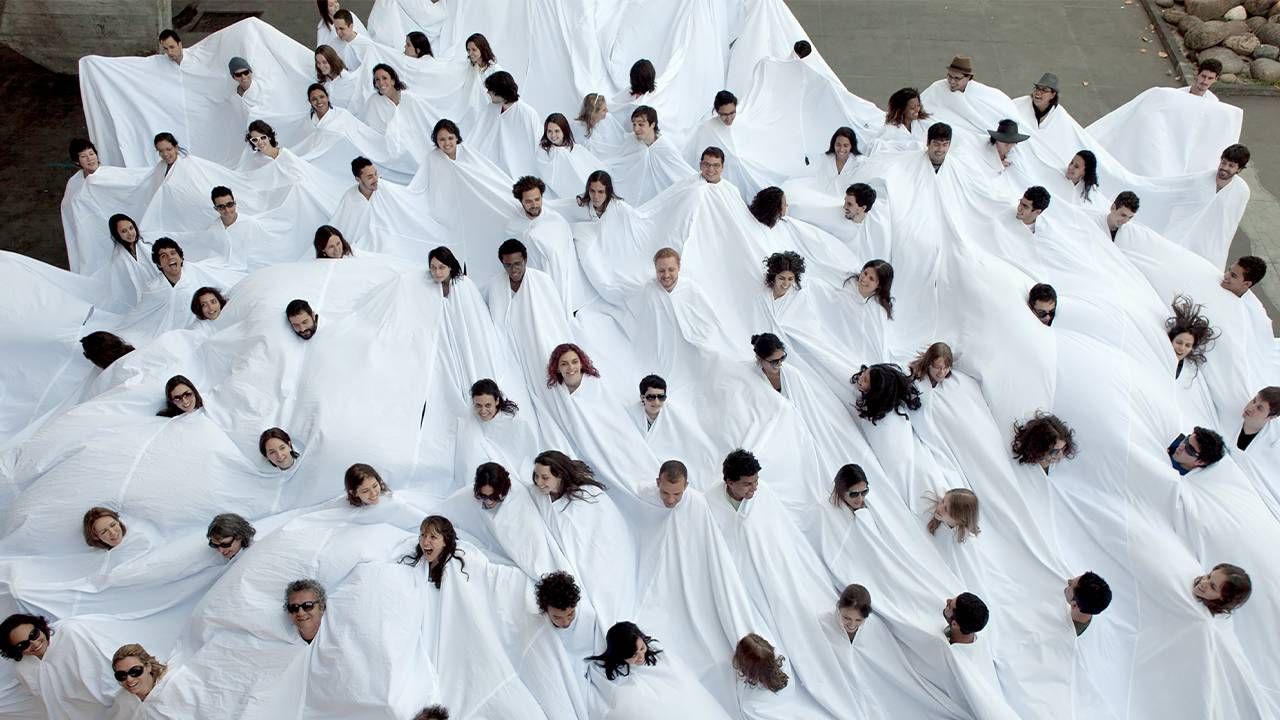 A group of people with their heads poking out of a giant white cloth. Next Avenue, art exhibition, Assembly Required