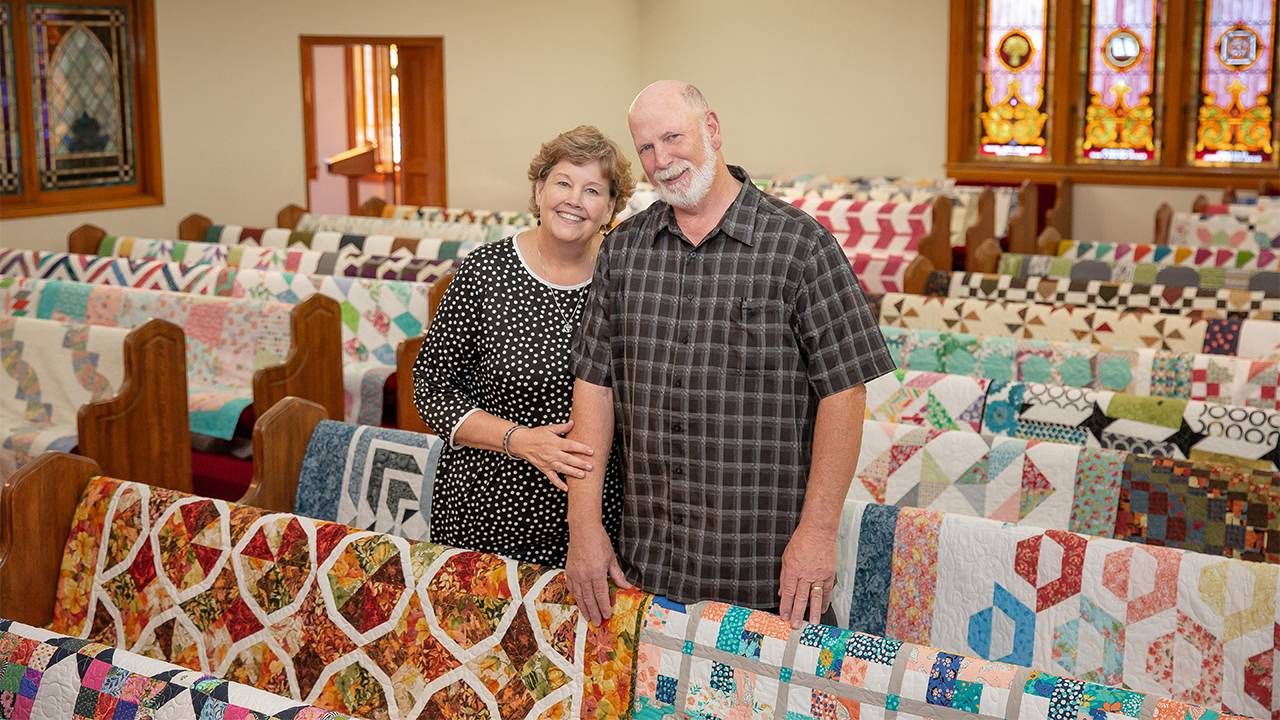An older couple smiling inside a church with colorful quilts laid over the pews. Next Avenue, quilting, Missouri Star Quilting Company MSQC