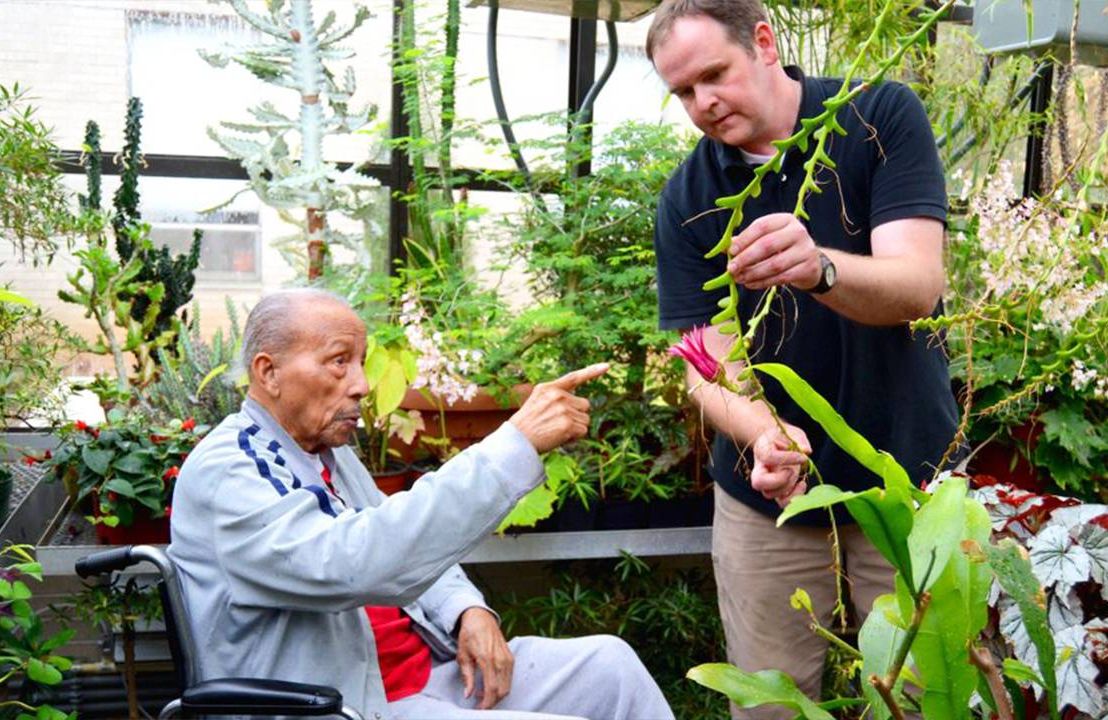 Two older men in a greenhouse, showing off the flowers. Next Avenue, benefits of flowers, mind and body