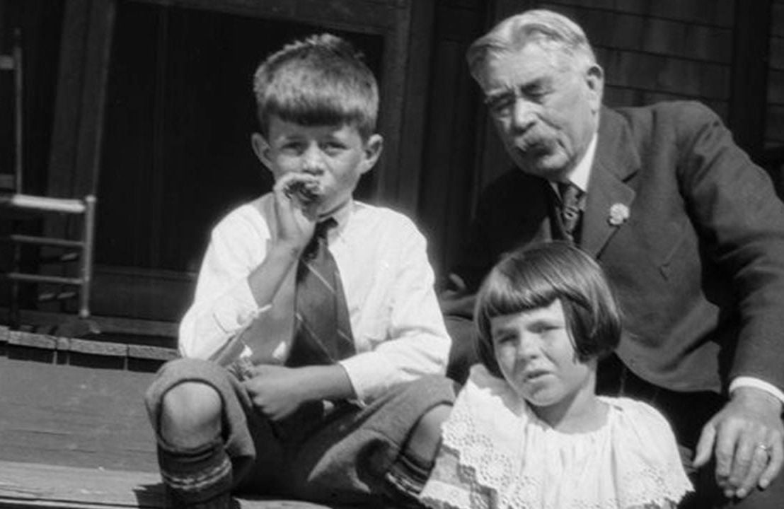 A black and white photo of two children and an older adult sitting on a stoop. Next Avenue, The First Kennedys