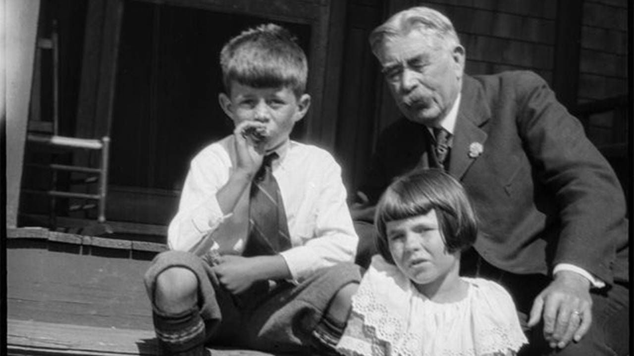 A black and white photo of two children and an older adult sitting on a stoop. Next Avenue, The First Kennedys