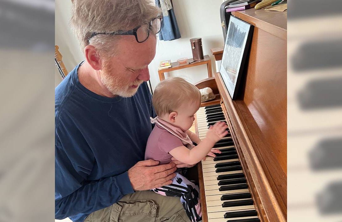 A grandparent with his granchild on his lap playing the piano. Next Avenue, grandparenting, grandparents child care