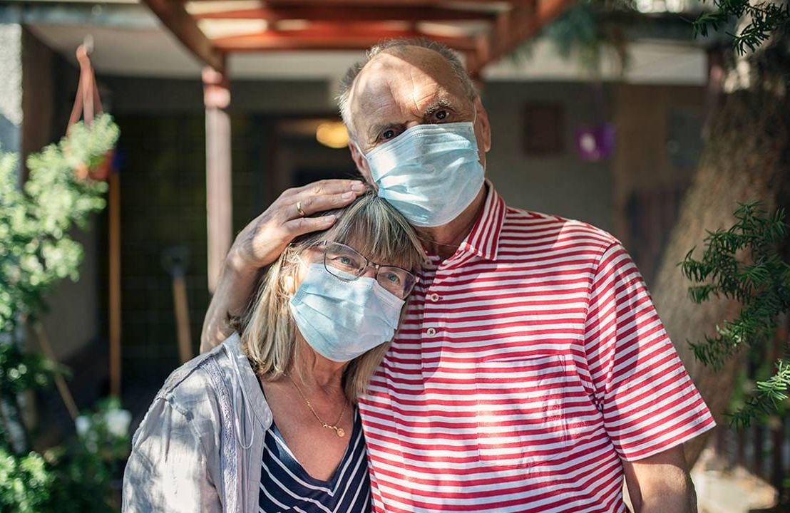 A couple standing outside their house wearing facemasks. Next Avenue, returning to normal, Immunocompromised, pandemic
