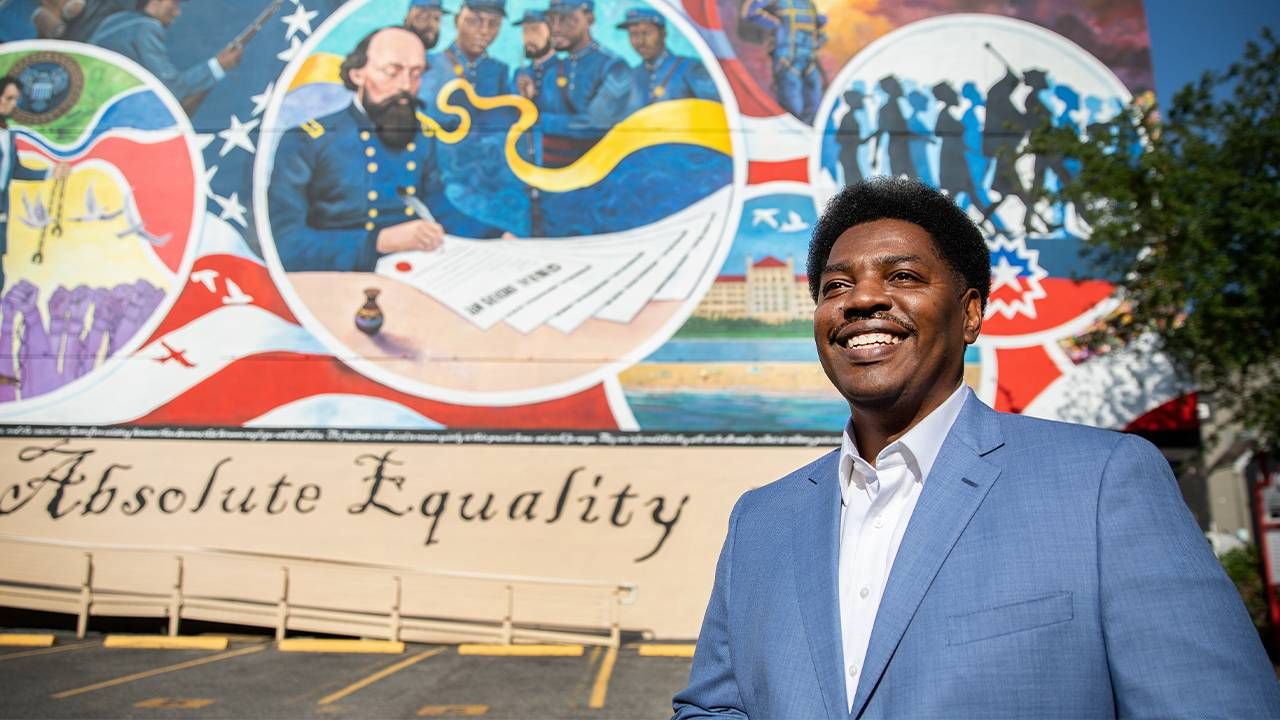 A close up of a man standing and smiling in front of a mural. Next Avenue, Galveston Juneteenth mural